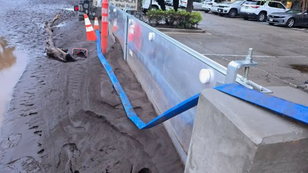 Flood barrier with metal panels installed along a muddy roadside, with a blue hose and concrete posts.